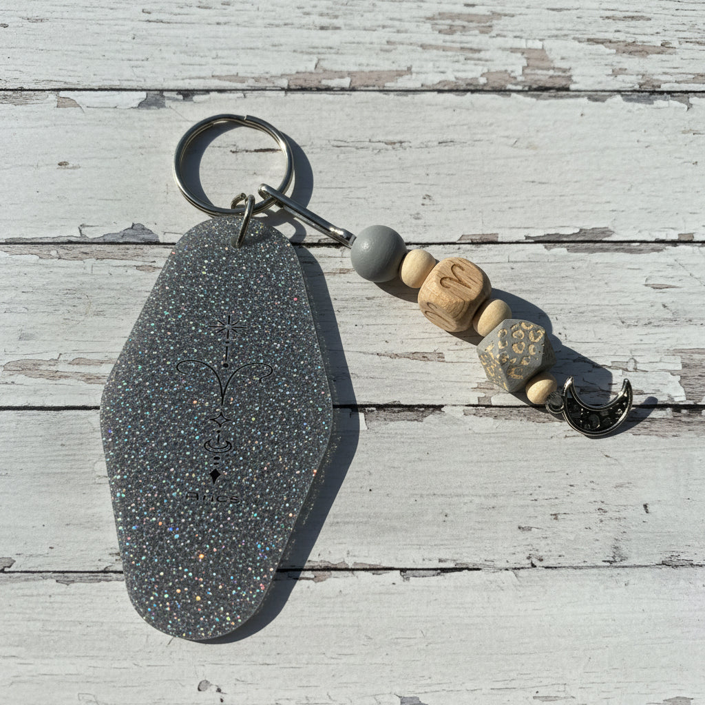 A handmade keychain featuring a large, glittery silver-gray teardrop-shaped charm engraved with a crescent moon and sun, attached to a metal split ring. The keychain also includes a gray bead, three wooden beads with carved letters (M, N, S), a patterned bead, and a small silver crescent moon charm, all displayed on a white background.