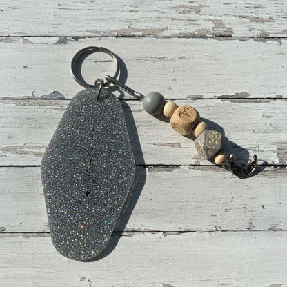 A handmade keychain featuring a large, glittery silver-gray teardrop-shaped charm engraved with a crescent moon and sun, attached to a metal split ring. The keychain also includes a gray bead, three wooden beads with carved letters (M, N, S), a patterned bead, and a small silver crescent moon charm, all displayed on a white background.