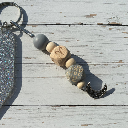 A close-up of a beaded keychain charm with wooden beads, a gray bead, a silver crescent moon charm, and a silver key ring, placed on a white background.