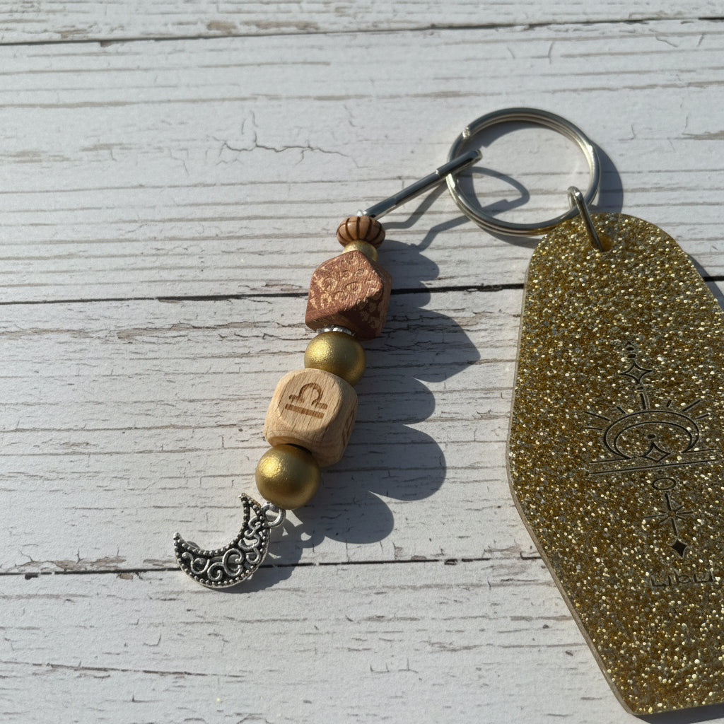 A beaded keychain with a gold glitter tag, a silver crescent moon charm, and wooden beads on a white background.