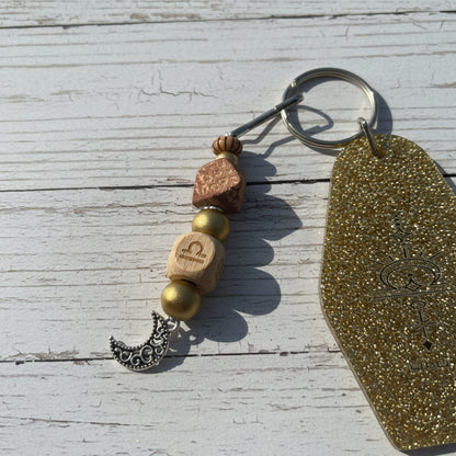 A beaded keychain with a gold glitter tag, a silver crescent moon charm, and wooden beads on a white background.