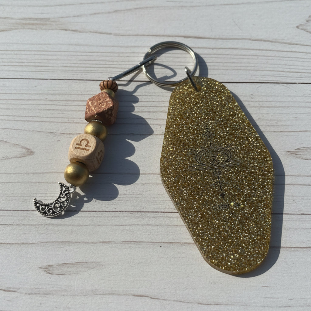 A bohemian-style keychain featuring a large, textured gold glitter tag with an engraved symbol, accompanied by wooden beads, a silver crescent moon charm, and a metal key ring, displayed on a white background.