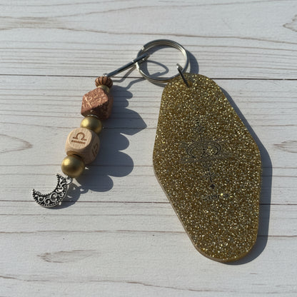 A bohemian-style keychain featuring a large, textured gold glitter tag with an engraved symbol, accompanied by wooden beads, a silver crescent moon charm, and a metal key ring, displayed on a white background.