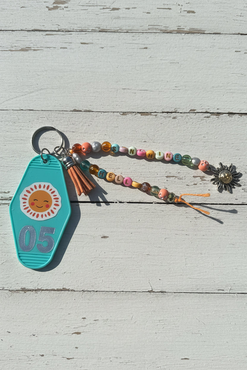 A turquoise plastic keychain with a smiling sun graphic and the number 05, attached to a beaded wristlet strap with colorful letter beads and a silver flower charm, resting on a white surface.