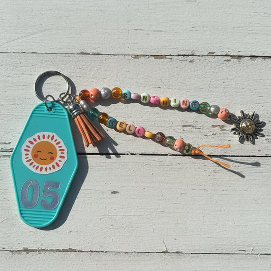 A turquoise plastic keychain with a smiling sun graphic and the number 05, attached to a beaded wristlet strap with colorful letter beads and a silver flower charm, resting on a white surface.