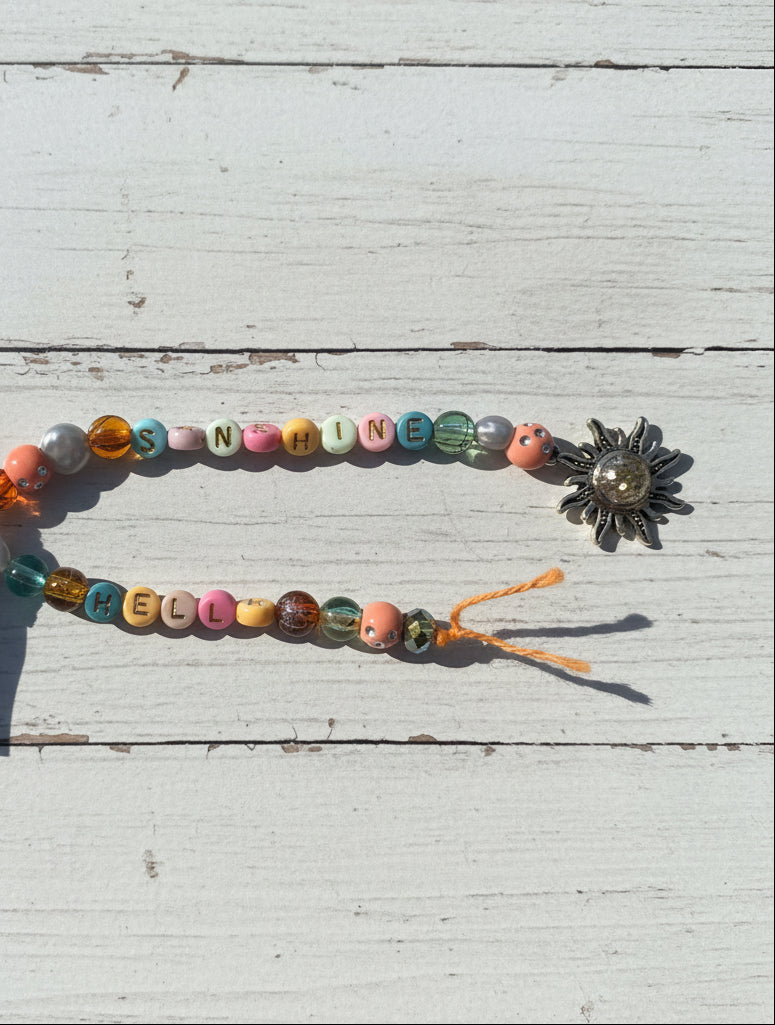 A handmade beaded necklace or bracelet laid on a brown kraft paper background. The top strand spells out 'SUNSHINE' using pastel-colored letter beads, while the bottom strand reads 'HELLO' with multicolored decorative beads.