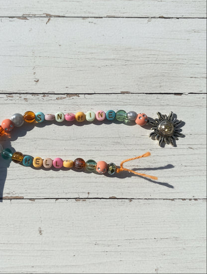 A handmade beaded necklace or bracelet laid on a brown kraft paper background. The top strand spells out 'SUNSHINE' using pastel-colored letter beads, while the bottom strand reads 'HELLO' with multicolored decorative beads.