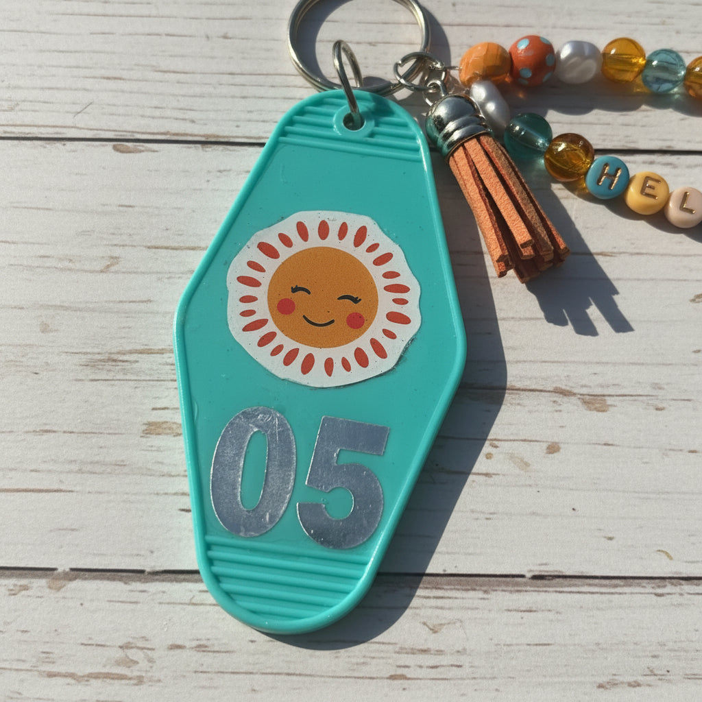 A turquoise plastic keychain or bag charm with a smiling sun sticker and the number 05 in silver, shown on a brown kraft paper background with part of a tassel keychain visible.