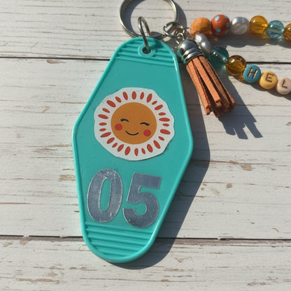 A turquoise plastic keychain or bag charm with a smiling sun sticker and the number 05 in silver, shown on a brown kraft paper background with part of a tassel keychain visible.