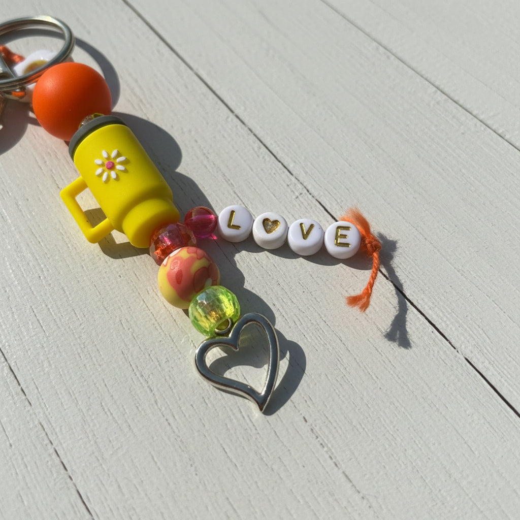 A beaded keychain charm laid on a tan paper background. The keychain includes a yellow mug bead with a white flower, translucent pink and green beads, white round beads spelling LOVE with gold lettering, a small orange heart bead, an orange tassel, and a silver heart-shaped metal charm.