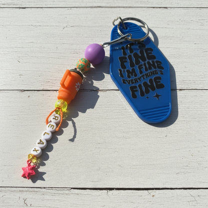 A beaded keychain with a blue flip-flop-shaped charm that reads 'IT'S FINE I'M FINE EVERYTHING'S FINE' in black text, alongside an orange whistle bead, white letter beads spelling 'RELAX', a purple bead, and colorful decorative accents, all on a white wooden background.