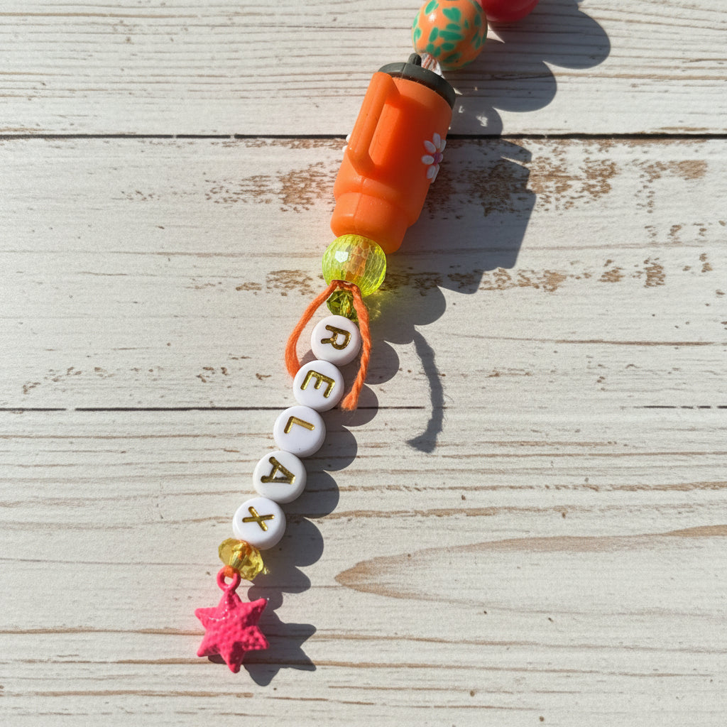 A close-up of a colorful beaded keychain or bag charm laid on a tan surface. The charm includes white round beads spelling 'RELAX' with gold lettering, an orange bottle-shaped section, a translucent green bead, a pink star charm, and small yellow glitter accents.