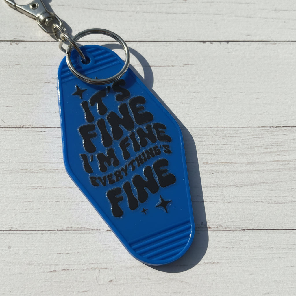 A bright blue keychain shaped like a flip-flop sandal with the text 'IT'S FINE I'M FINE EVERYTHING'S FINE' in black lettering, attached to a silver metal key ring, set against a whitebackground.