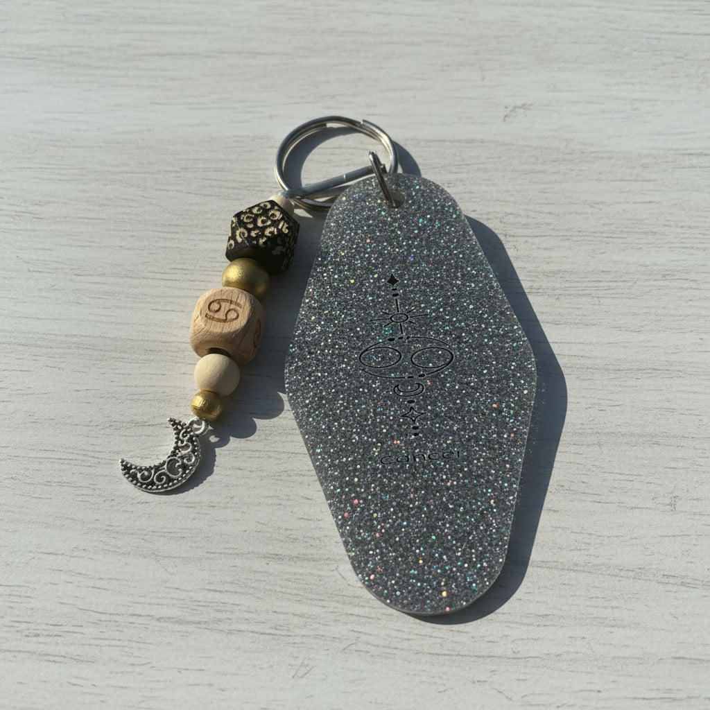 A silver-toned keychain with a large, glittery hexagonal charm featuring a black chakra symbol, accented by wooden beads, a black filigree bead, and a silver crescent moon charm, displayed on a brown kraft paper background.