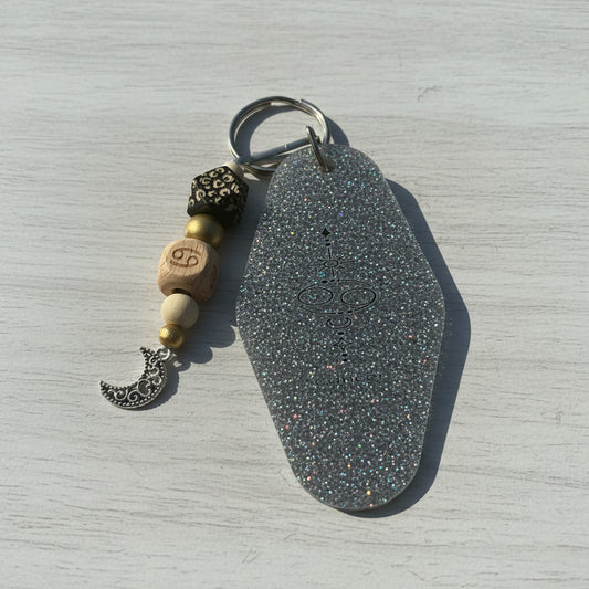 A silver-toned keychain with a large, glittery hexagonal charm featuring a black chakra symbol, accented by wooden beads, a black filigree bead, and a silver crescent moon charm, displayed on a brown kraft paper background.