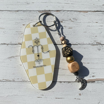 A zodiac-themed keychain featuring a coffin-shaped tag with a beige and white checkerboard pattern and a Scorpio symbol, alongside decorative beads and a silver crescent moon charm, displayed on a white background.