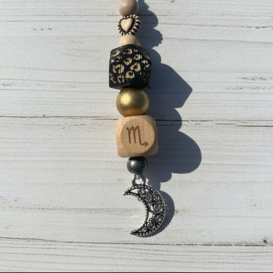 A close-up of a beaded charm or necklace with a silver crescent moon charm, a wooden bead engraved with a gold bead, and other decorative beads on a white background.