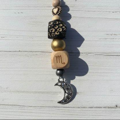A close-up of a beaded charm or necklace with a silver crescent moon charm, a wooden bead engraved with a gold bead, and other decorative beads on a white background.