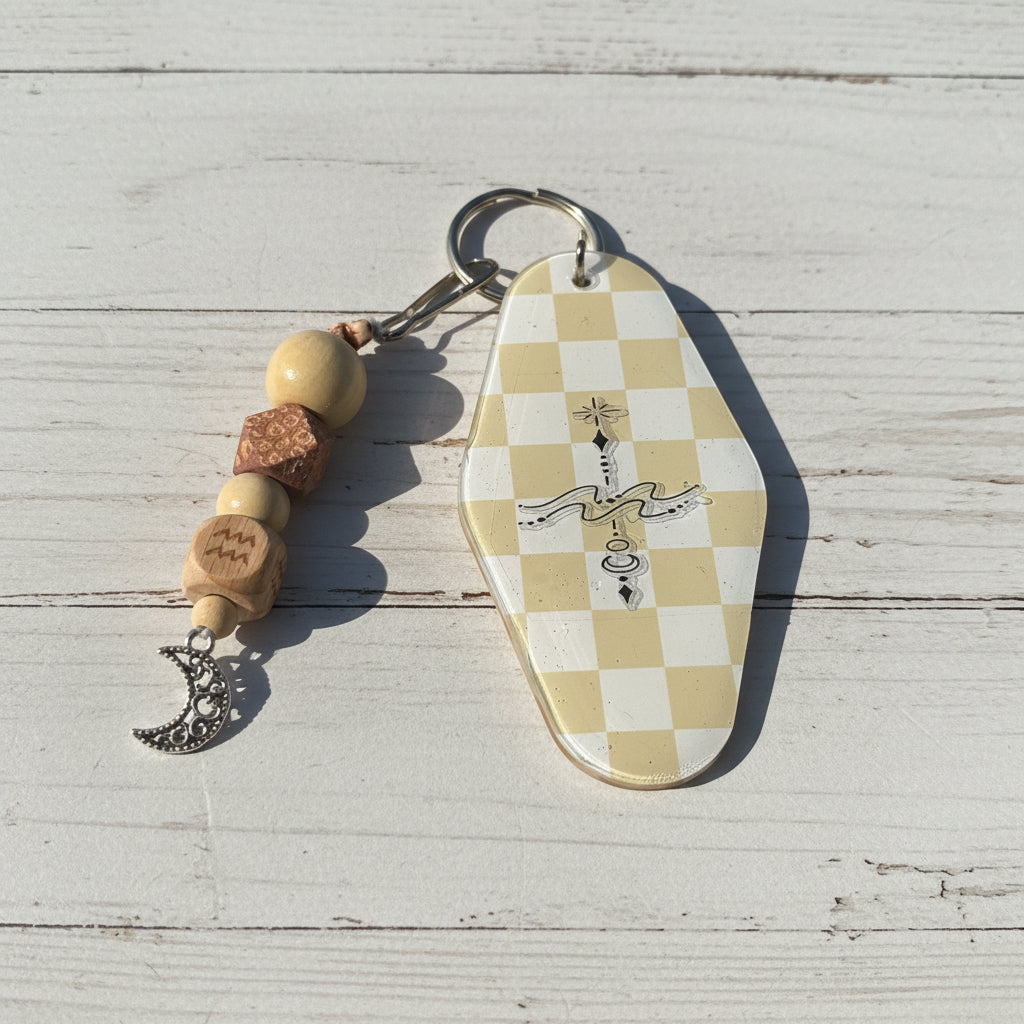 A coffin-shaped keychain with a cream and beige checkerboard pattern and a black illustration of a key and ribbon, attached to a beaded keychain with wooden beads and a silver crescent moon charm, set against a white background.