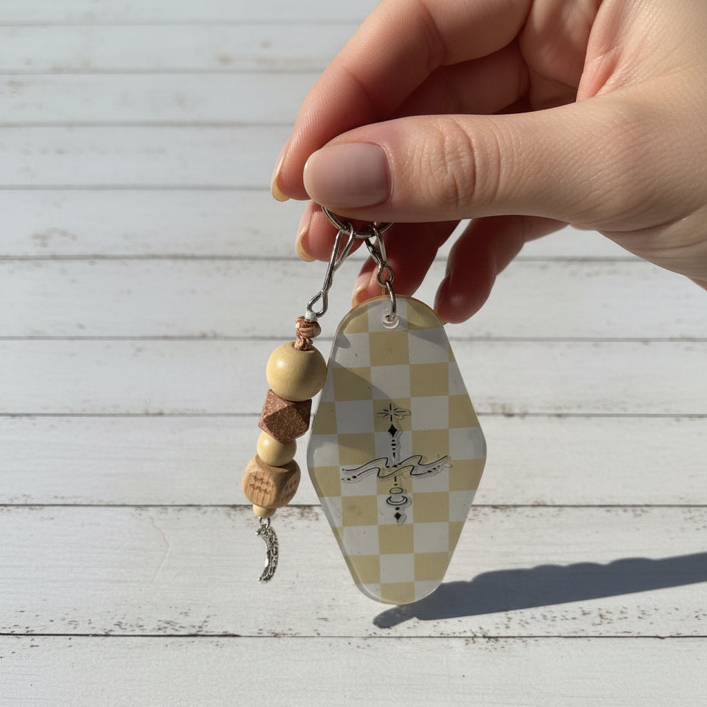 A beaded keychain charm with light wood beads, a small pink patterned cube, a silver crescent moon charm, and a cream-and-beige checkered key tag with black line art, photographed on a white background.