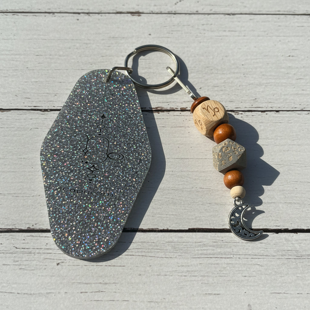 A glittery keychain with an irregular silver stone-shaped tag, a split key ring, wooden beads, a gray geometric bead, a white bead, and a silver crescent moon charm, displayed on a white background.