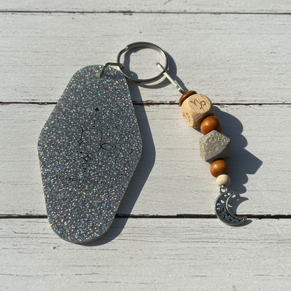 A glittery keychain with an irregular silver stone-shaped tag, a split key ring, wooden beads, a gray geometric bead, a white bead, and a silver crescent moon charm, displayed on a white background.