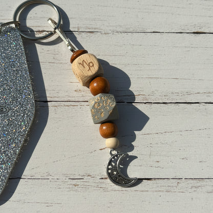 A close-up of a beaded keychain with a silver crescent moon charm, resting on a brown kraft paper background. The keychain features a silver keyring, a silver-toned wooden cube with an engraved zodiac symbol, orange-brown wooden beads, a textured gray cube, a small white bead, and the silver crescent moon charm.