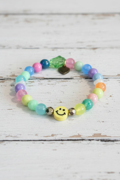 A colorful beaded bracelet with pastel round beads, a green faceted bead, a small gold heart charm, and a yellow smiley face charm