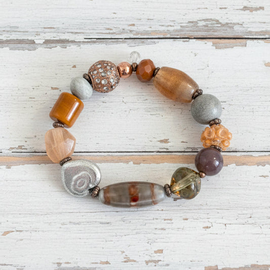 A beaded stretch bracelet with a mix of earthy, metallic, and stone-like beads in brown, orange, silver, and green.
