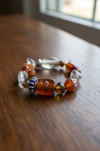A beaded stretch bracelet featuring various translucent and opaque beads in brown, orange, clear, and silver, with a prominent carved brown bead at the center