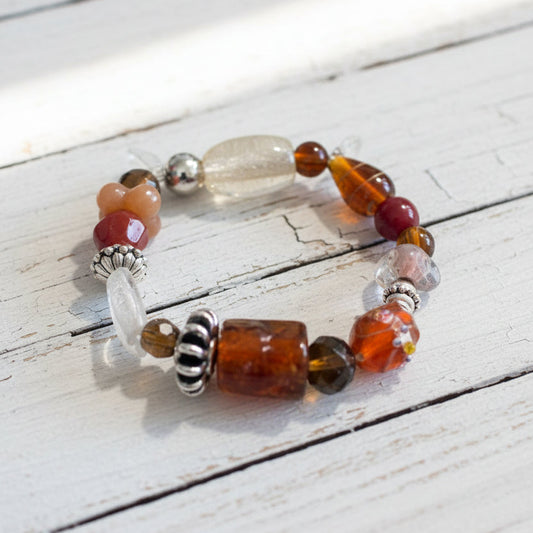 A beaded stretch bracelet featuring a large central brown cylindrical bead, a clear faceted bead, and smaller red, orange, and silver beads.