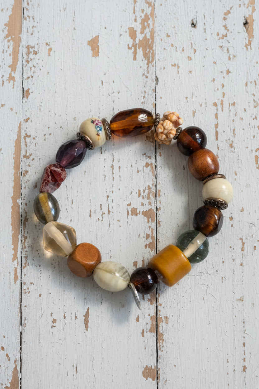 A beaded stretch bracelet featuring a mix of polished and textured stone beads in brown, orange, white, and black, with a small silver metal charm, displayed on a white background.