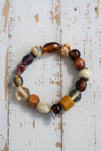 A beaded stretch bracelet featuring a mix of polished and textured stone beads in brown, orange, white, and black, with a small silver metal charm, displayed on a white background.