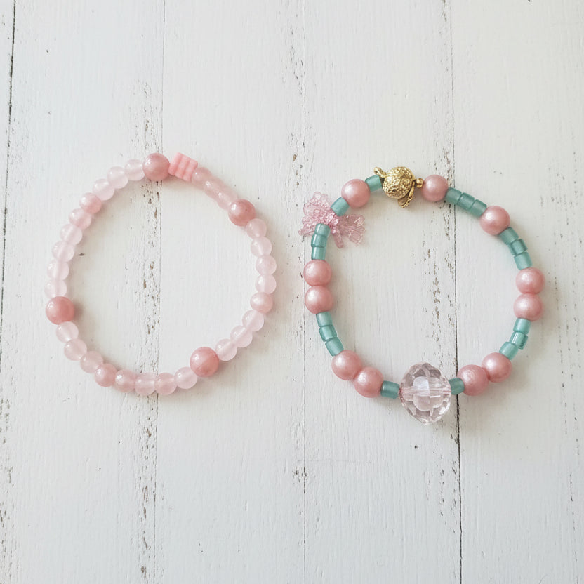 Two beaded bracelets displayed on a white wood. One bracelet is made of light pink beads, and the other is made of turquoise blue beads with a central pink heart charm.