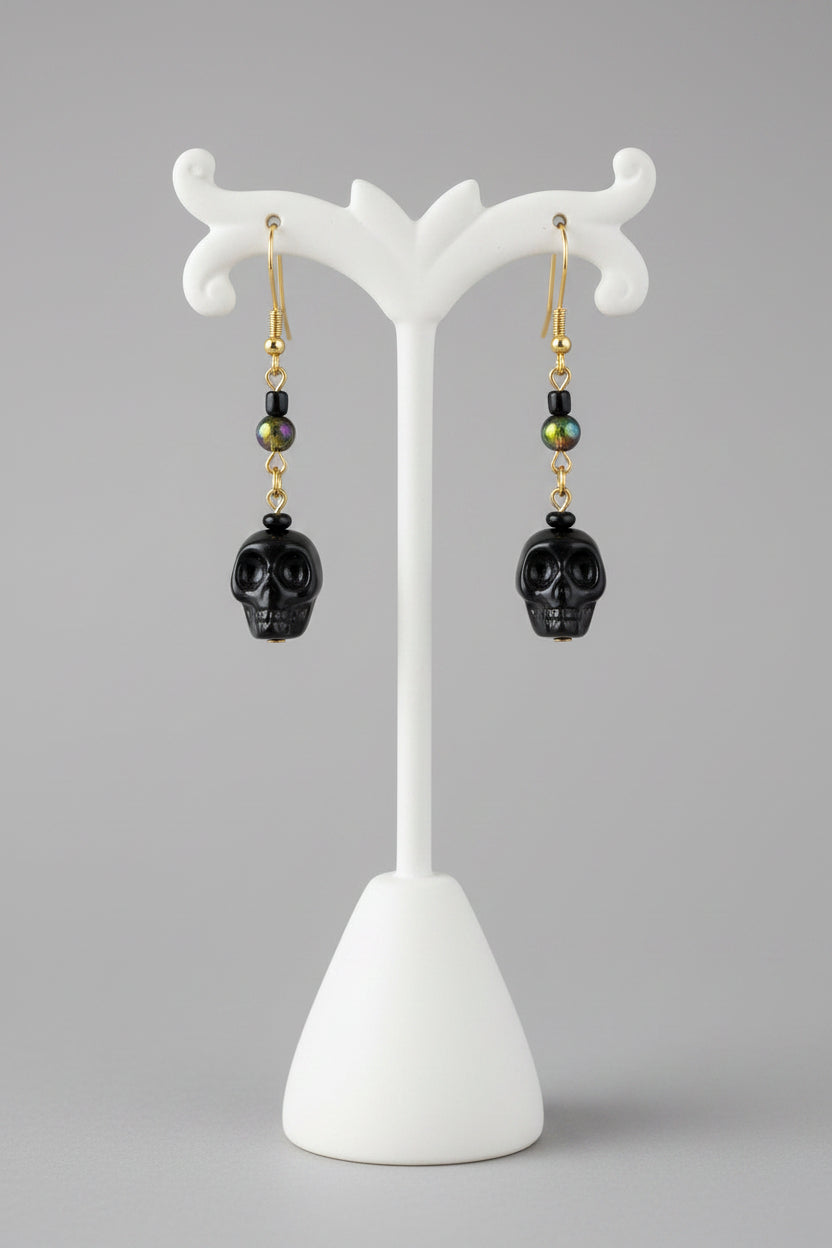 Black skull drop earrings with gold and green bead accents, hanging from a wooden display stand.