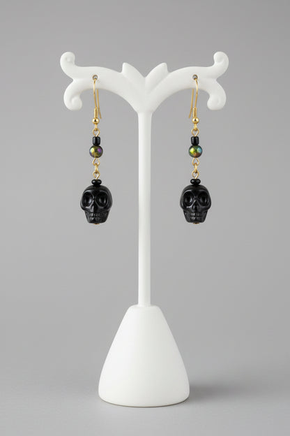 Black skull drop earrings with gold and green bead accents, hanging from a wooden display stand.