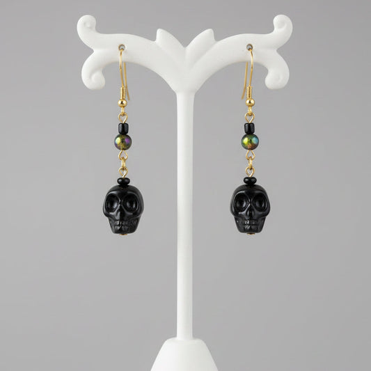 Black skull drop earrings with gold and green bead accents, hanging from a wooden display stand.