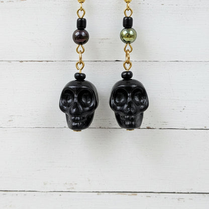 Black skull drop earrings with gold chain links and dark beads, hanging in front of a white backdrop.