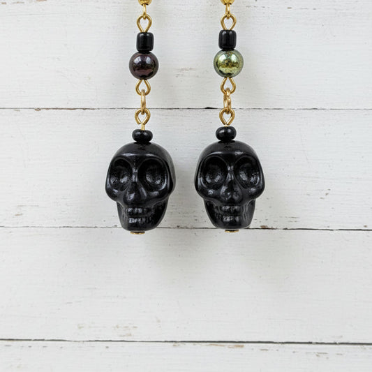 Black skull drop earrings with gold chain links and dark beads, hanging in front of a white backdrop.