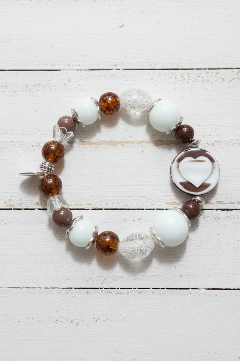 A beaded stretch bracelet featuring white, brown, and clear beads, including a prominent white heart-shaped bead, displayed on a white background.