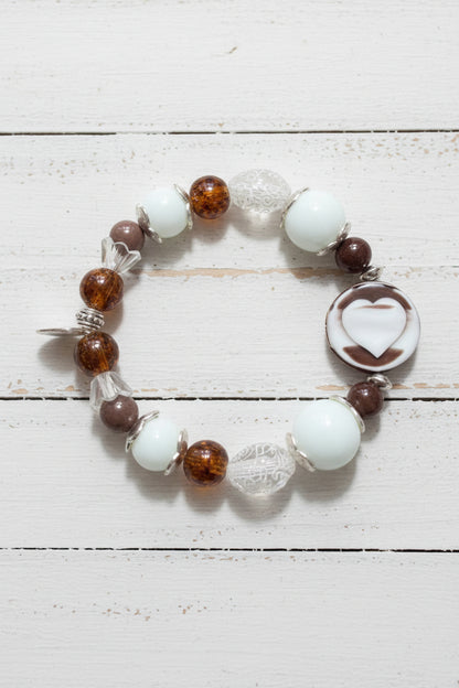 A beaded stretch bracelet featuring white, brown, and clear beads, including a prominent white heart-shaped bead, displayed on a white background.