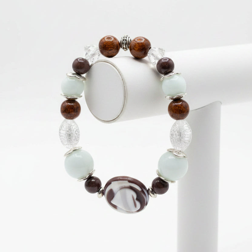 A beaded stretch bracelet featuring light blue-green turquoise beads, dark brown translucent beads, a central white and black marbled oval charm, and silver-toned spacer beads, displayed on a stand