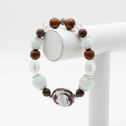 A beaded stretch bracelet featuring light blue-green turquoise beads, dark brown translucent beads, a central white and black marbled oval charm, and silver-toned spacer beads, displayed on a stand