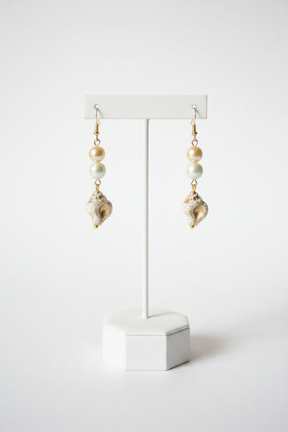 A pair of dangling earrings with gold-tone hooks, light blue beads, white pearl-like beads, and textured white and gold shell charms, displayed on a stand.