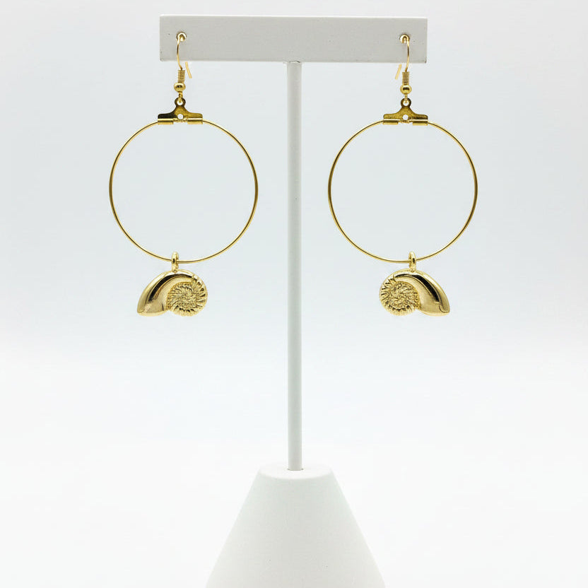 A pair of gold hoop earrings with textured shell charms, displayed on a earring stand