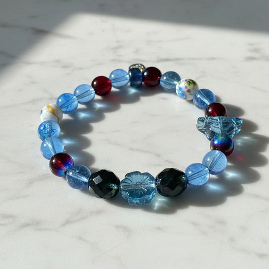 A beaded stretch bracelet featuring faceted blue, dark blue, and red beads, along with a dark blue whale tail charm, displayed on a granite background