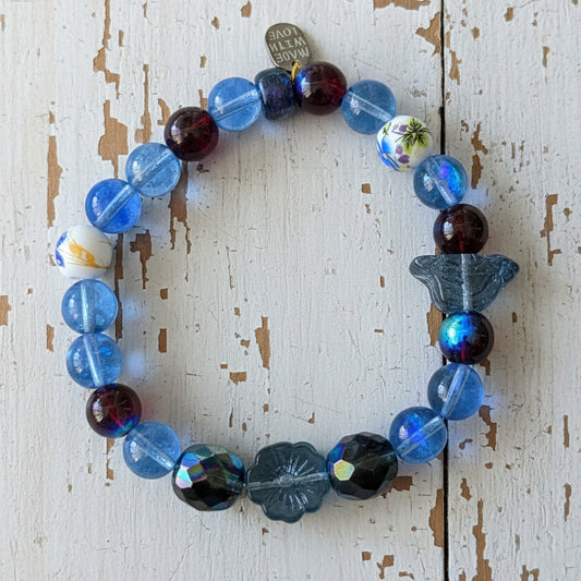 A beaded stretch bracelet featuring blue, dark blue, and red translucent beads, a butterfly-shaped bead, and a silver-toned tag, displayed on a wihite background.