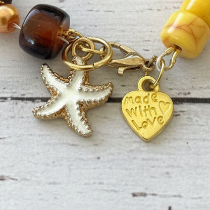 A close-up of a beaded charm bracelet featuring a white starfish charm, a gold heart charm engraved with 'Made with Love', and amber-colored beads on a white textured background.