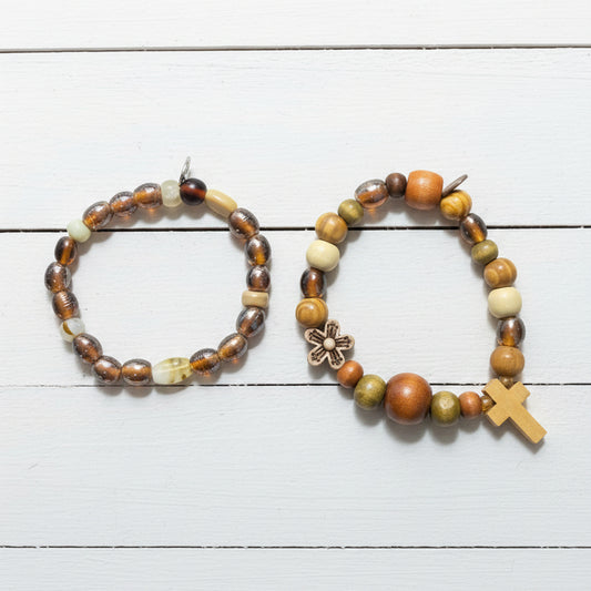 Two beaded bracelets displayed on a brown paper bag. One bracelet features brown and black marbled beads with a small silver-toned cross charm, while the other features earthy brown, green, and cream beads with a small wooden cross charm.
