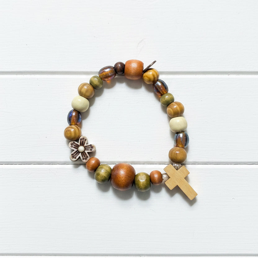 A beaded stretch bracelet featuring a mix of earthy brown, orange, olive green, and cream beads, a small white flower charm, and a light wood cross, displayed on a white background.